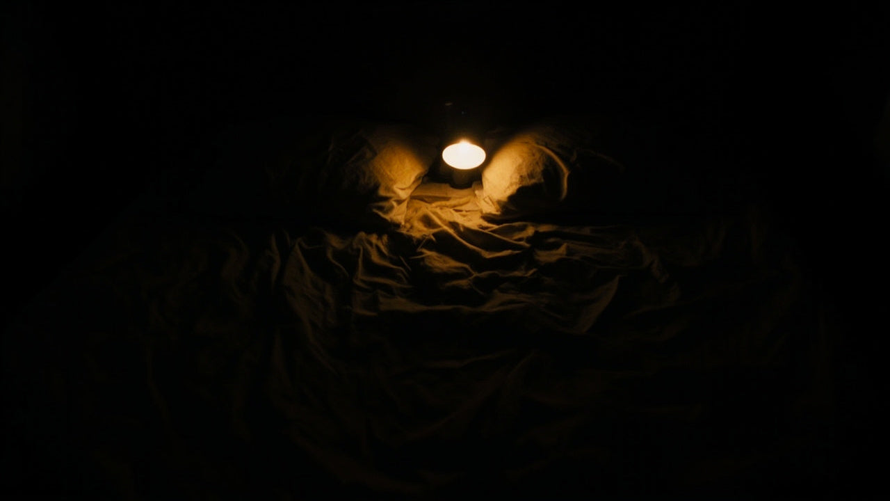 A dimly lit bedroom at night, a single lamp glowing between two pillows on a rumpled, empty bed.