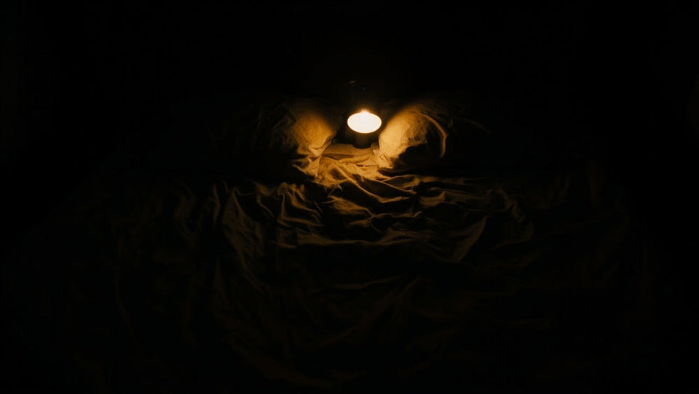 A dimly lit bedroom at night, a single lamp glowing between two pillows on a rumpled, empty bed.